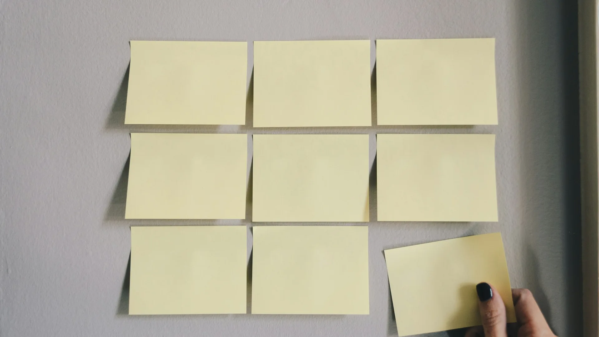 Nine blank yellow sticky notes on a wall, with a hand placing one more note