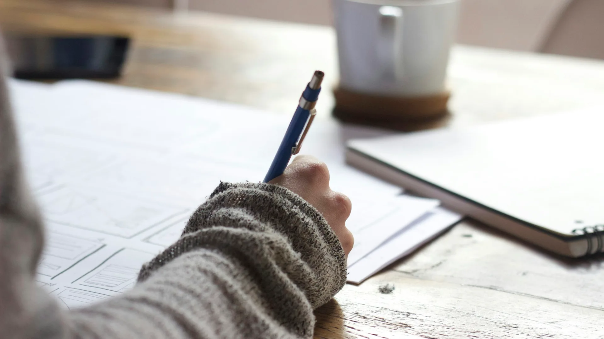Person writing on paper at a wooden table with a notebook nearby