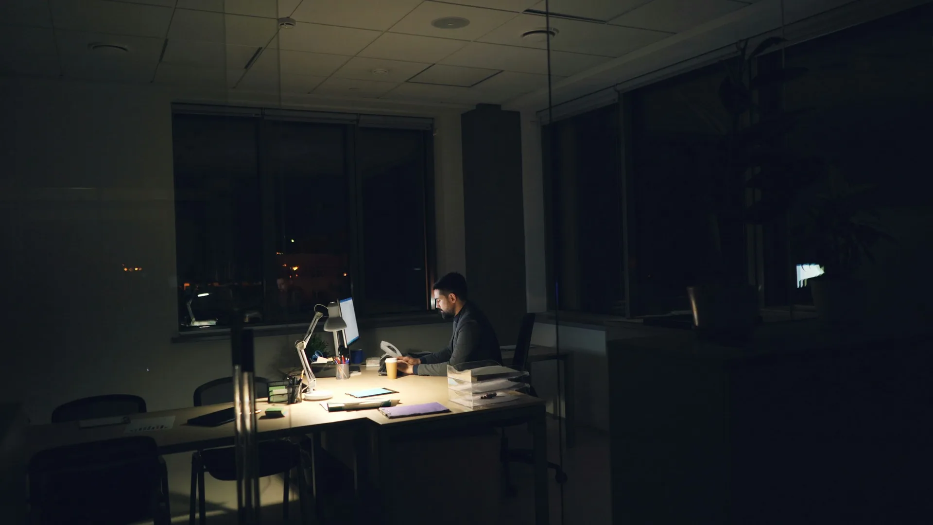 Person working alone in a dark office at night, lit by a desk lamp and a computer screen