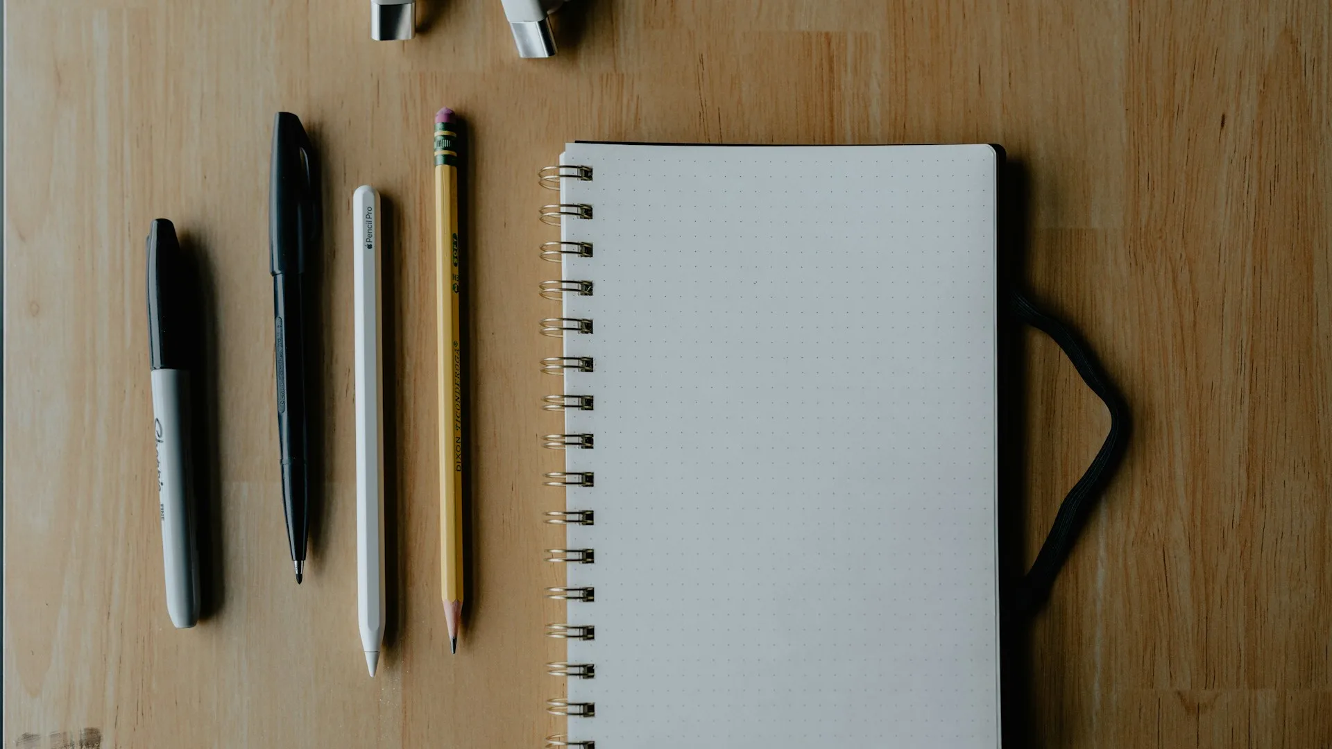 Four pens and pencils lined up beside an open notebook on a wooden desk